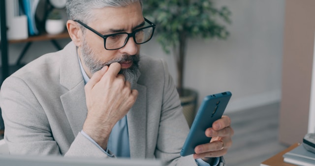 Ein Mann sitzt am Schreibtisch und blickt auf ein Smartphone.